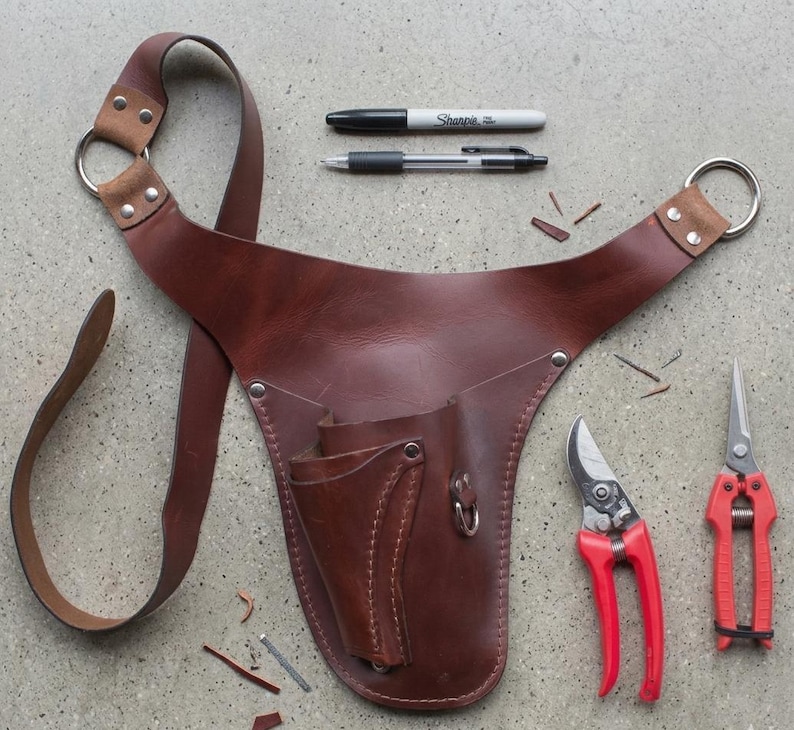 May include: A brown leather gardening tool belt with a shoulder strap, two metal rings, and multiple pockets. Two pairs of red handled pruning shears and two pens are also visible.