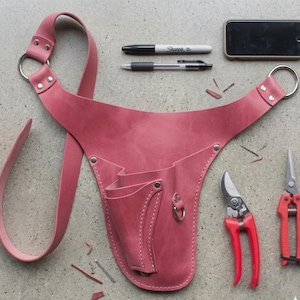 May include: A dusty rose leather gardening tool belt with multiple pockets and a shoulder strap. Two pairs of red handled pruning shears, a black pen, a black Sharpie, and a smartphone are also present.