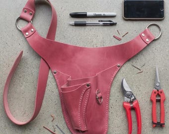 May include: A dusty rose leather gardening tool belt with multiple pockets and a shoulder strap. Two pairs of red handled pruning shears, a black pen, a black Sharpie, and a smartphone are also present.