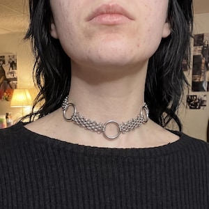 Puede incluir: Un collar tipo gargantilla de cota de malla en tono plateado que presenta tres grandes anillos metálicos circulares conectados por segmentos de eslabones metálicos tejidos intrincados. El diseño es simétrico, con un anillo central y dos secciones.