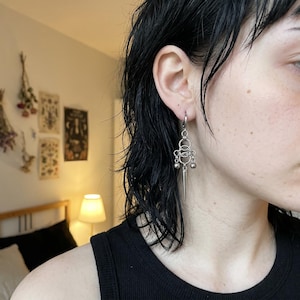 Peut inclure: Une boucle d'oreille pendante en métal argenté composée d'une attache en anneau reliée à une série de cercles entrelacés, deux petites perles sphériques et une pointe centrale dirigée vers le bas suspendue au bas de la structure.