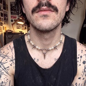 Peut inclure: Un collier ras-du-cou composé d'un rang de perles sphériques blanches alternées avec des pointes en métal argenté. Le collier présente un anneau central en métal argenté avec un pendentif en forme de pointe dirigé vers le bas.