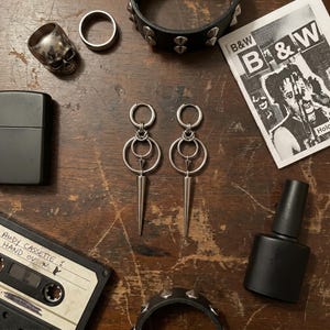 Könnte beinhalten: Eine Sammlung alternativer Accessoires auf einer Holzoberfläche. Enthält silberne Ohrringe, Ringe, einen Totenkopfring, ein schwarzes Feuerzeug, eine Kassette, eine schwarze Nagellackflasche und Armbänder mit Nieten. Ein Schwarz-Weiß-Zine ist ebenfalls vorhanden.