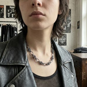 Op de afbeelding: Een zilverkleurige ketting met een ingewikkeld ontwerp van verbonden ovale en bloemachtige elementen. De ketting wordt gedragen over een zwarte mesh top en een zwarte leren jas.