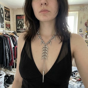 Puede incluir: Collar de metal plateado con un diseño de colgante de púas. El collar tiene una cadena con múltiples púas y un colgante central más largo con púas. La joya se lleva sobre un top negro.
