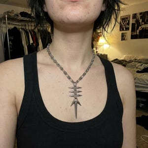 Op de afbeelding: Zilverkleurige ketting met een schakelkettingontwerp en een hanger met een reeks spikes en een centraal pijlvormig element. De ketting wordt gedragen over een zwarte tanktop.