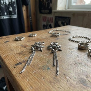 Könnte beinhalten: Eine Sammlung von Silberschmuck, darunter Ringe, Armbänder und Ohrringe, die auf einer Holzoberfläche ausgestellt sind. Die Ohrringe haben ein Sternmuster mit hängenden Ketten. Die Ringe haben verschiedene Designs.