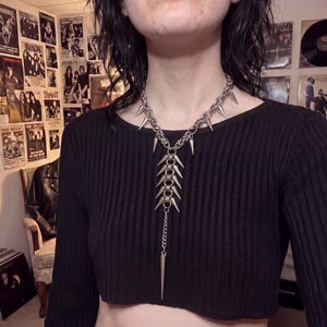 Puede incluir: Un collar de cadena de metal plateado con una serie de púas puntiagudas unidas a anillos entrelazados. El diseño incluye una sección central vertical con múltiples púas y una púa larga que cuelga de una cadena fina en la parte inferior.