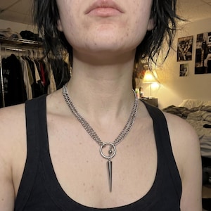 Op de afbeelding: Zilverkleurige ketting met een dubbel kettingontwerp. Een grote ring verbindt de kettingen, met een hangende, langwerpige kegelvormige hanger. De ketting wordt gedragen over een zwarte tanktop.