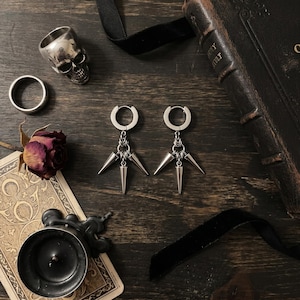 Op de afbeelding: Zilveren oorringen met drie kegelvormige hangers. De afbeelding toont ook een zilveren schedelring, een eenvoudige zilveren ring, een gedroogde roos en een vintage boek op een donker houten oppervlak.