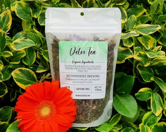 May include: A white pouch with a green label that says "Detox Tea" and lists the organic ingredients. The pouch contains a blend of dried herbs. The pouch is sitting on a bed of green and yellow leaves.