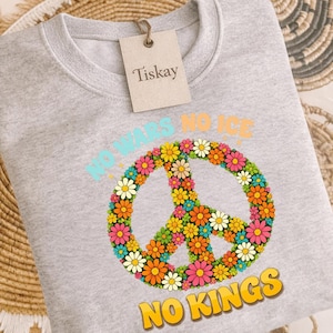 Może przedstawiać: Jasnoszara bluza z nadrukiem kwiatowego znaku pokoju. Znak pokoju wykonany jest z kolorowych kwiatów. Na bluzie nadrukowano tekst "NO WARS NO ICE" i "NO KINGS".