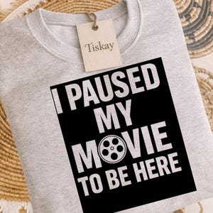 Puede incluir: Sudadera gris claro con el texto blanco "I PAUSED MY MOVIE TO BE HERE" sobre un rectángulo negro. La etiqueta de la marca dice "Tiskay". Es una prenda de vestir informal.