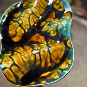 May include: An irregular, organic-shaped ceramic dish featuring a glossy glaze with a marbled pattern of deep black, golden yellow, and vibrant teal. The edges are finished in a solid teal, creating a distinct contrast against the dark interior.