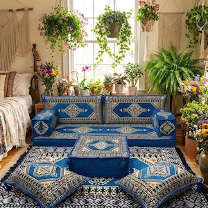 May include: A bohemian-style living space featuring a low, blue patterned sofa and matching cushions. The room is decorated with plants, flowers, and macrame wall hangings, creating a cozy and inviting atmosphere. The rug and cushions have a geometric design.