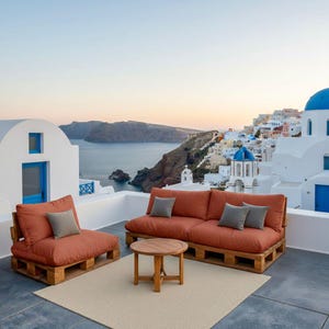 May include: Outdoor patio furniture set featuring a rust-colored sofa and armchair with gray accent pillows. The furniture sits on wooden pallets, with a small round wooden table and a beige rug. The background shows a scenic view of a coastal town.