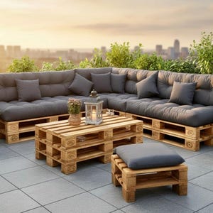 May include: Outdoor sectional sofa and coffee table made from wooden pallets. The sofa has dark gray cushions and pillows. A small footstool with a matching cushion is also present. The setting is on a patio with potted plants and a city skyline in the background.