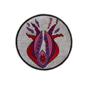 May include: A round patch with a silver background and a black border. The patch features a stylized floral design in shades of red, pink, and purple. The design has a central flower shape.