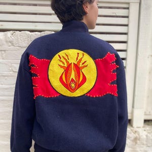 May include: A navy blue bomber jacket with a high collar and a large, colourful emblem on the back. The emblem features a yellow circle with a red and orange design, flanked by red patches with decorative stitching.