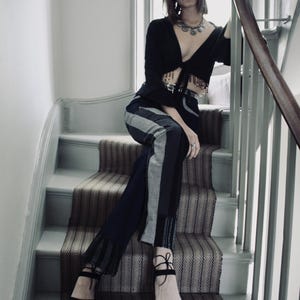 May include: A woman's ensemble comprising a black tie-front top, high-waisted trousers with vertical stripes, and black platform sandals with red soles. The trousers have a patchwork design. The woman is seated on a staircase with a patterned carpet.