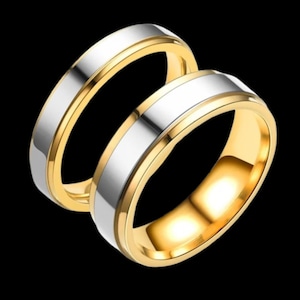 Könnte beinhalten: Zwei Eheringe mit einem goldenen Band und einem silbernen Mittelband werden vor einem schwarzen Hintergrund gezeigt. Die Ringe liegen eng beieinander und zeigen ihre kontrastierenden Farben und die polierte Oberfläche. Das Gold reflektiert das Licht.