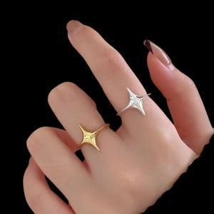 以下が含まれることがあります： 金と銀の2つのリング。それぞれ4つの尖った星のデザインが特徴です。黒い背景に、手の上にリングが展示されています。スターバーストのデザインは、モダンでミニマリストなスタイルです。