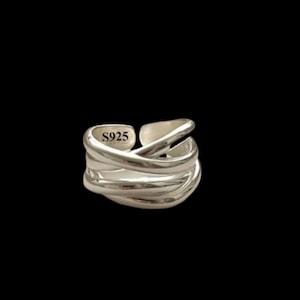May include: A silver-colored ring with a layered, crisscross design. The ring has an adjustable open band and features the text "S925" on the inside. The ring is set against a black background.