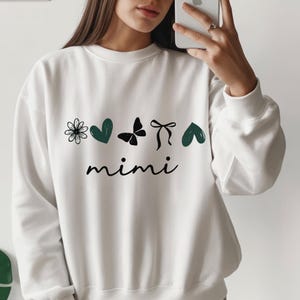Könnte beinhalten: Weißes Sweatshirt mit floralem Design, grünen Herzen, einem Schmetterling, einer Schleife und dem Wort "mimi" in schwarzer Schrift. Ein lässiges Oberteil für den Alltag.