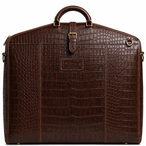 Könnte beinhalten: Eine dunkelbraune Kleidersacktasche mit einer strukturierten Krokodilleder-Optik. Die Tasche verfügt über einen Tragegriff, einen vertikalen Mittelriemen mit goldfarbener Schnalle und goldfarbene Metallbeschläge an den Seiten für einen Schultergurt.