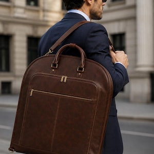 K&ouml;nnte beinhalten: Eine dunkelbraune Kleidersacktasche mit glatter Oberfl&auml;che, ausgestattet mit einem Tragegriff, einem langen verstellbaren Schulterriemen und einer gro&szlig;en rechteckigen Vordertasche mit goldfarbenen Metallrei&szlig;verschl&uuml;ssen. Die Tasche hat eine abgerundete Oberkante.