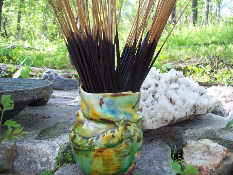 May include: A bundle of incense sticks in a colorful ceramic vase. The vase has a green, yellow, and blue glaze. The incense sticks are brown and have a natural wood scent.