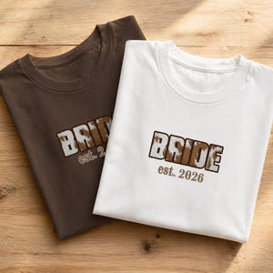 Könnte beinhalten: Zwei T-Shirts, eines braun und eines weiß, werden auf einer Holzoberfläche gezeigt. Jedes Shirt hat das Wort "BRIDE" in einem Blockbuchstabenstil mit einem Kuhfellmuster und den Text "est. 2026" darunter. Die Shirts sind gefaltet.