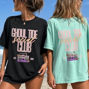 May include: A black t-shirt and a light green t-shirt, each featuring the text "GHOUL TIDE social CLUB" in pink and tan, with a graphic of a purple boombox decorated with skulls and flowers, and the text "EST. 2026" printed on the front.
