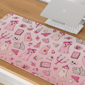 May include: A rectangular desk mat featuring a repeating pattern on a light pink background. The design includes illustrations of ghosts wearing witch hats, books labeled Book of Spells, skulls, mushrooms, candles, potion bottles, and candy.