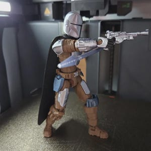 May include: A Mandalorian action figure in a dynamic pose, holding a blaster pistol and a spear. The figure is clad in silver and brown armor with a black cape. The figure is approximately 6 inches tall.