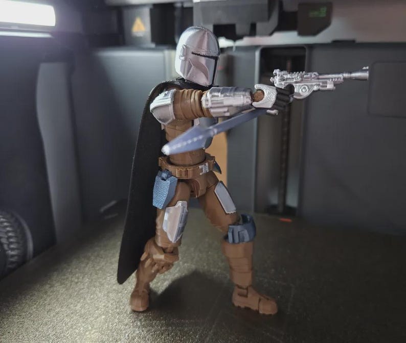 May include: A Mandalorian action figure in a dynamic pose, holding a blaster pistol and a spear. The figure is clad in silver and brown armor with a black cape. The figure is approximately 6 inches tall.