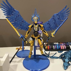 May include: A detailed action figure with blue wings, gold armor, and two gold swords. The figure stands on a blue circular base. The wings are spread wide, and the figure has a helmet with a cross design. The figure is a fantasy-themed collectible.