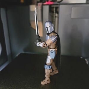 May include: A Mandalorian action figure in a brown and silver suit, holding a black and silver sword. The figure has a black cape and is posed in a dynamic stance. The figure is on a dark surface.
