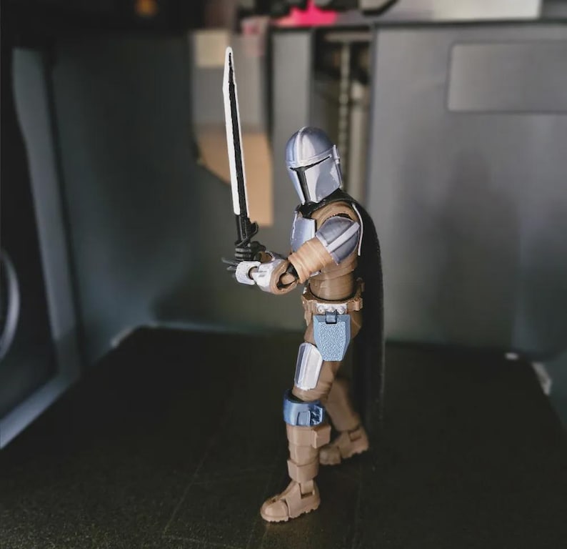 May include: A Mandalorian action figure in a brown and silver suit, holding a black and silver sword. The figure has a black cape and is posed in a dynamic stance. The figure is on a dark surface.