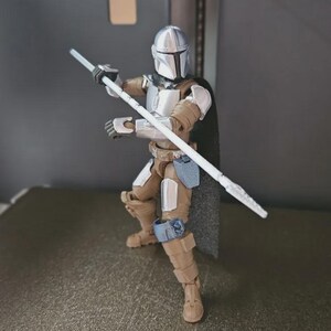 May include: A Mandalorian action figure in a dynamic pose, holding a white staff. The figure is clad in silver and tan armor with a black cape. The figure is posed on a gray surface.