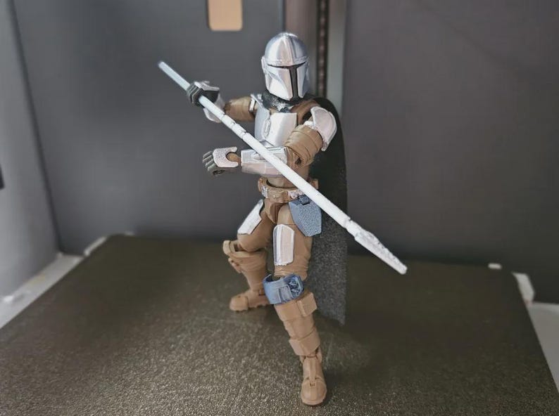 May include: A Mandalorian action figure in a dynamic pose, holding a white staff. The figure is clad in silver and tan armor with a black cape. The figure is posed on a gray surface.