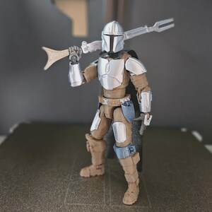 May include: A detailed action figure of a Mandalorian warrior, featuring a silver helmet and armor, with brown and tan clothing. The figure is holding a spear-like weapon over its shoulder and a smaller weapon in its right hand. The figure is standing on a dark surface.