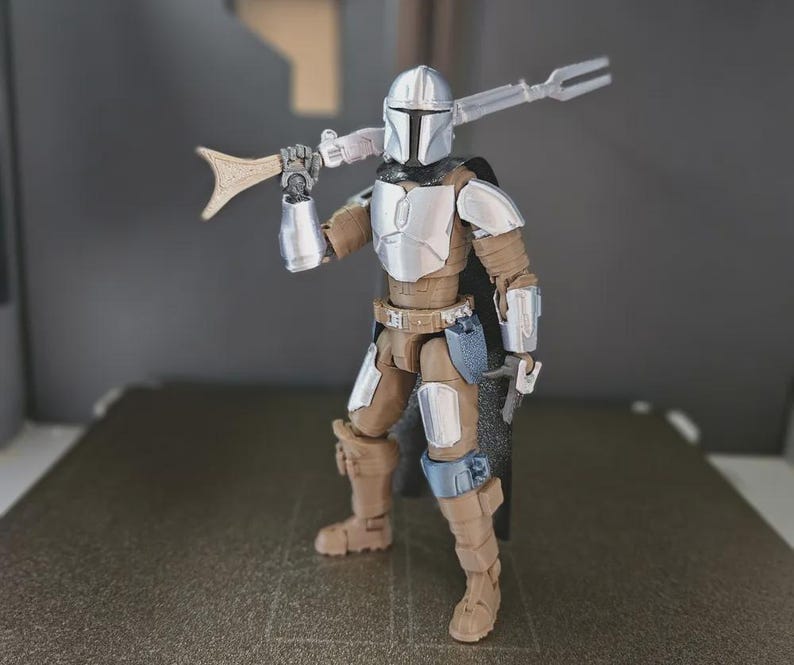 May include: A detailed action figure of a Mandalorian warrior, featuring a silver helmet and armor, with brown and tan clothing. The figure is holding a spear-like weapon over its shoulder and a smaller weapon in its right hand. The figure is standing on a dark surface.