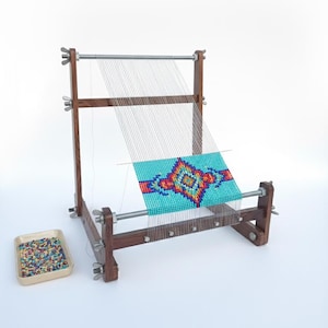 May include: A wooden bead loom with a partially completed beaded design in turquoise, red, orange, and yellow. The loom has a rectangular frame and adjustable metal hardware. A small tray of colourful beads sits to the side.