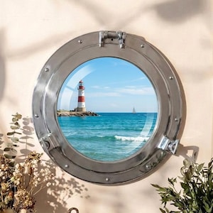 Puede incluir: Un espejo de ojo de buey redondo y plateado con una vista de un faro a rayas rojas y blancas en una isla rocosa. El océano y un velero son visibles a través del cristal. Hay objetos decorativos en primer plano.