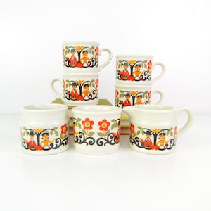 May include: Set of six white ceramic mugs with a colourful folk art design featuring red flowers, green leaves, and black silhouettes of people. The mugs have a rounded shape and a handle on the side. There is also a matching creamer.