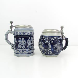 May include: Two ceramic beer steins with metal lids. One is light blue with a relief of figures, the other is dark blue with floral and figure relief. Both have handles and are on a white surface.