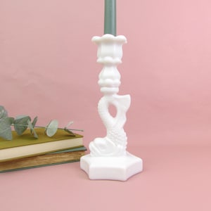 May include: A white ceramic candlestick holder with a fish design. The candlestick has a square base and an ornate body. A green candle is in the holder. The background is a solid pink colour.