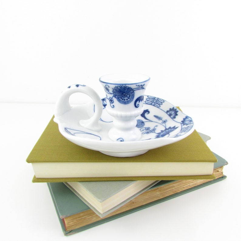 May include: A white porcelain candle holder with a blue and white floral design. The holder has a handle and a small base. It is sitting on top of a stack of books.
