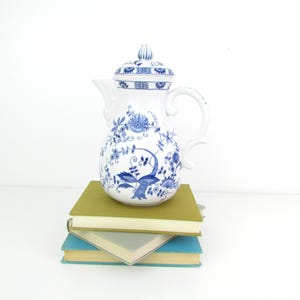 May include: A white porcelain teapot with a blue and white floral pattern. The teapot has a lid and a handle. It is sitting on top of a stack of books.